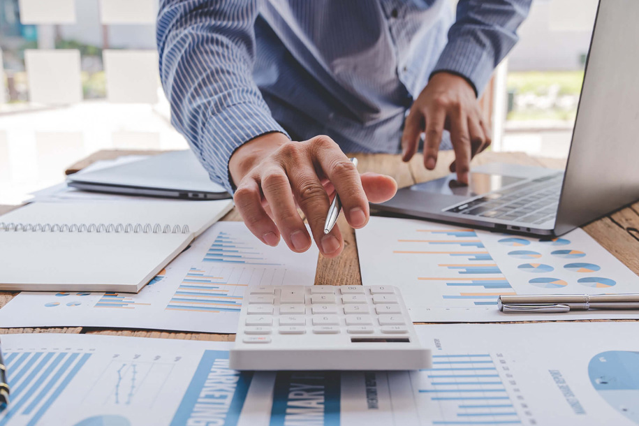 A person working at a desk using a calculator, laptop, and printed financial charts.